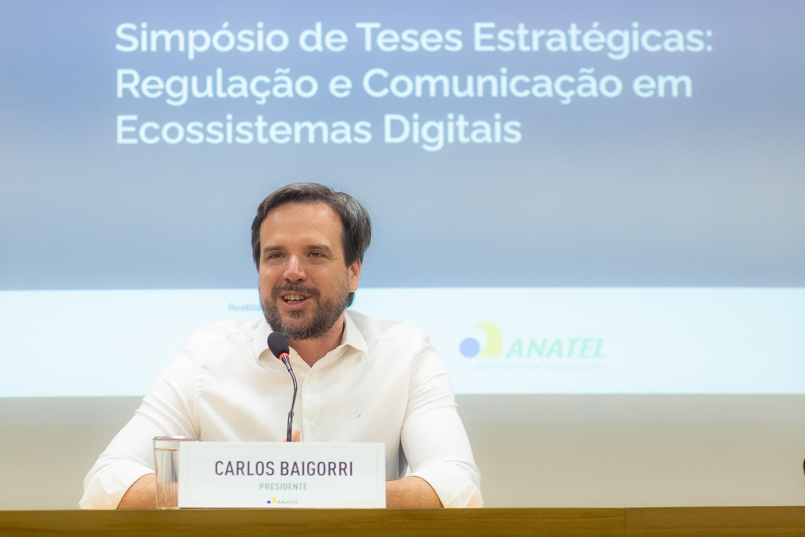 Presidente da ANATEL em fala na mesa do simpósio, com o telão ao fundo