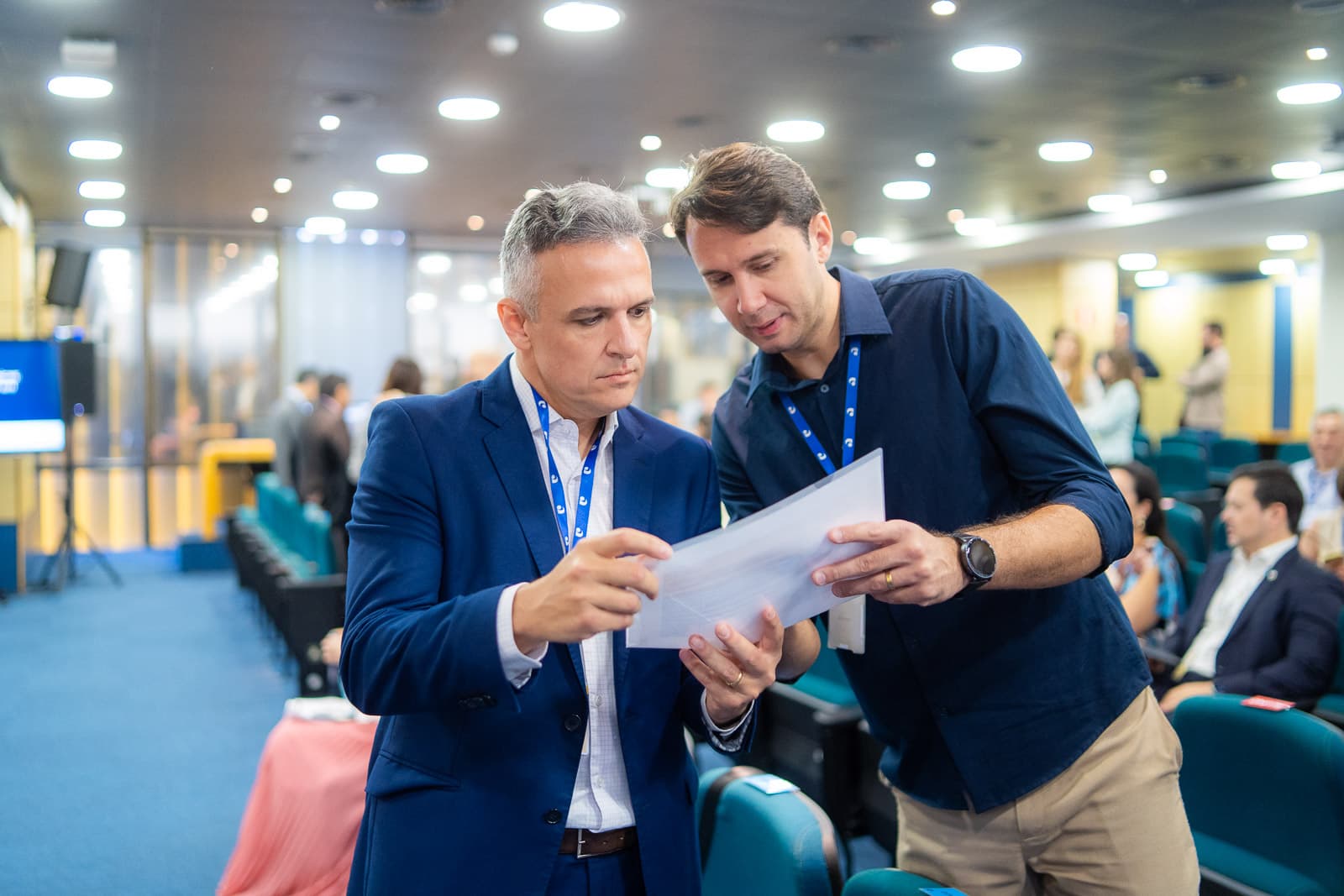 Dois participantes de um evento corporativo, com crachás, conferem um documento no corredor do auditório enquanto a plateia se acomoda ao fundo